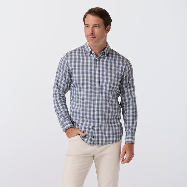 A man modeling a long-sleeve button-down shirt with a blue and white gingham pattern, paired with beige trousers, against a plain light gray studio background.